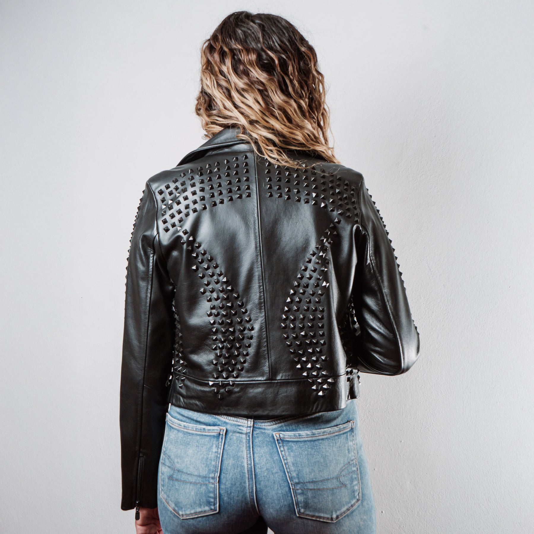 Mujer usando chamarra biker de piel negra con estoperoles negros modelo 'Rockstar Oscura' de Gerhel. Vista trasera.